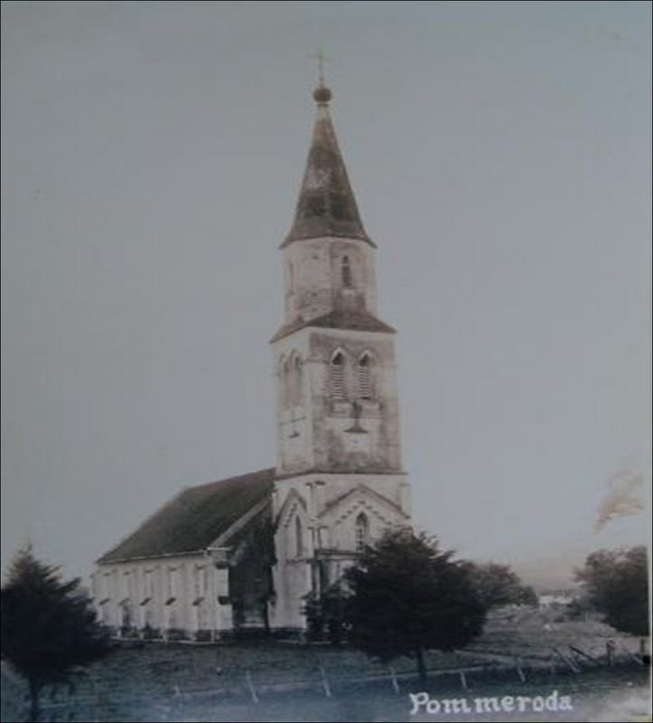 Imagem em preto e branco mostra uma igreja antiga com uma torre alta e fina, finalizada com um cume pontiagudo. A igreja é cercada por algumas árvores e parece estar situada em uma área rural ou em um campo aberto. A foto é visivelmente antiga, com qualidade típica de fotografias antigas em preto e branco.